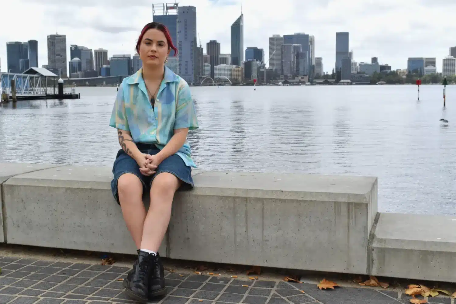 Finlaey Hewlett sits on a bench in front of the swan river. They are wearing a blue top and denim shorts.